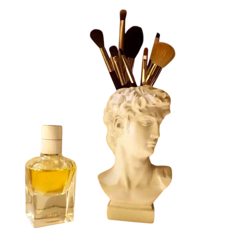 By Levin white resin David bust vase holding makeup brushes on a vanity next to a perfume bottle